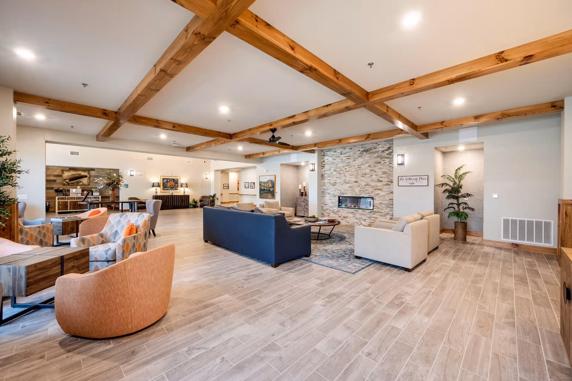 Spacious senior living facility common area with wooden ceiling beams, a modern electric fireplace set in a stone wall, multiple seating arrangements including sofas and armchairs, wooden flooring, and decorative plants and artwork.
