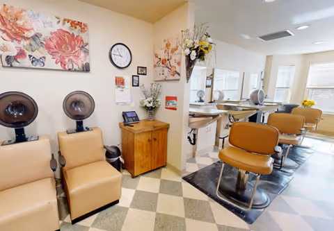Interior view of a salon area with beige salon chairs and hair drying stations on the left side. On the right side, there are salon styling chairs in front of mirrors and windows letting in natural light. The walls are decorated with floral paintings and a clock. A small wooden cabinet with a phone and flower arrangement is positioned in the corner.