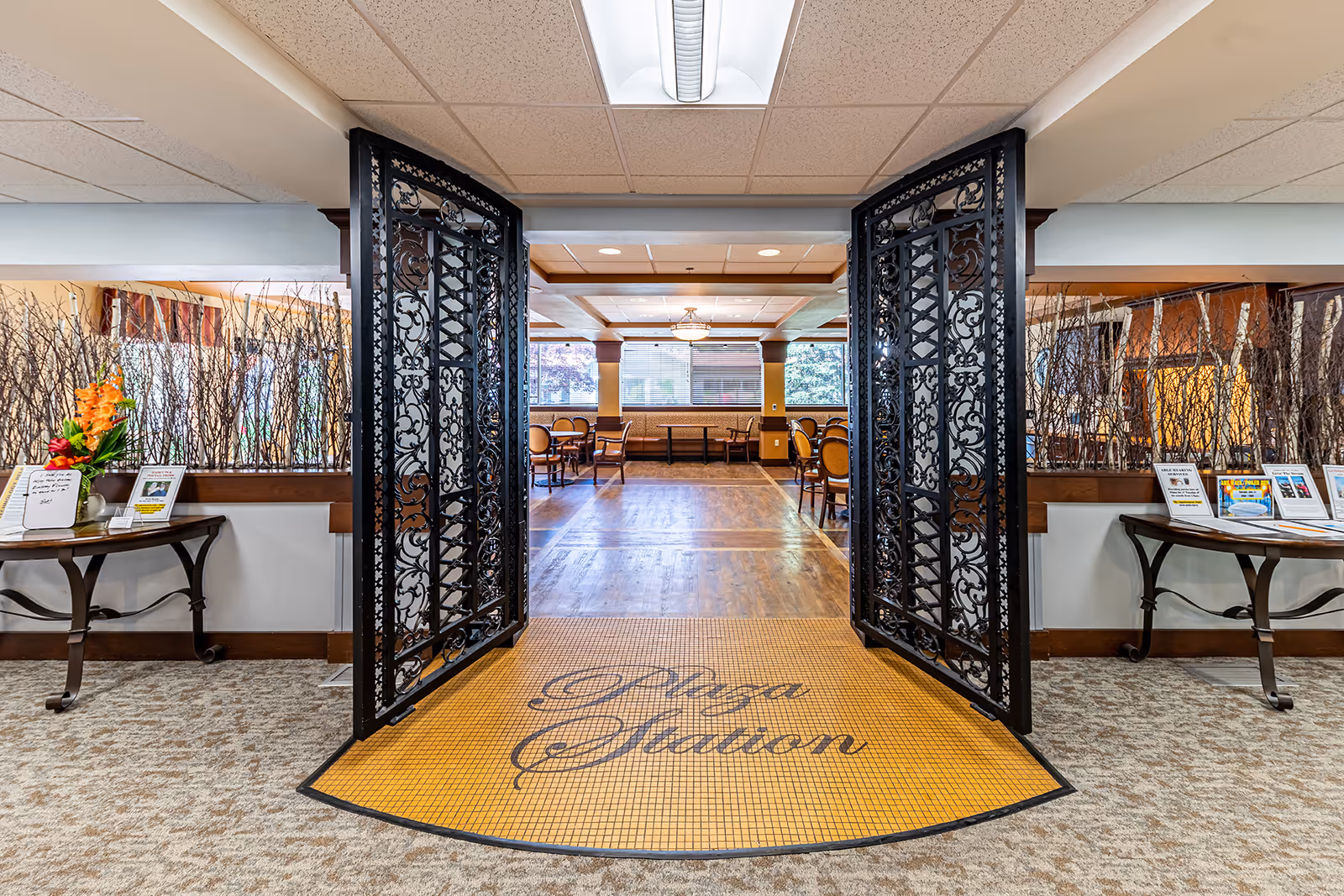 Entrance to a dining area named 'Plaza Station' with ornate black metal double doors open wide. The floor at the entrance has a yellow tiled mat with the words 'Plaza Station' written in cursive. Inside, there are wooden floors, tables, and chairs arranged neatly. On either side of the entrance, there are tables with decorative branches and informational flyers. The ceiling has recessed lighting and a central chandelier.