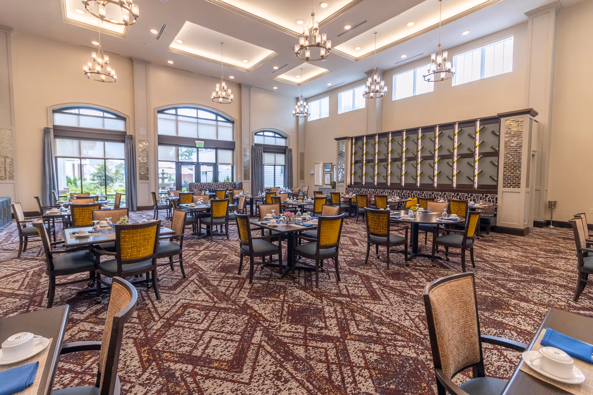 Spacious, well-lit dining room with multiple set tables and chairs, chandeliers, high windows, and a decorative wall display.