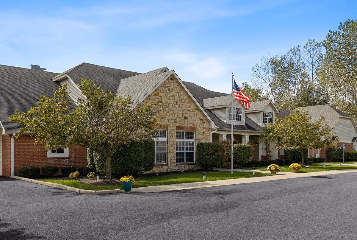 Exterior view of a single-story senior living facility building with a stone and brick facade, a neatly maintained lawn, small trees, and an American flag on a flagpole in front. The sky is clear and blue.