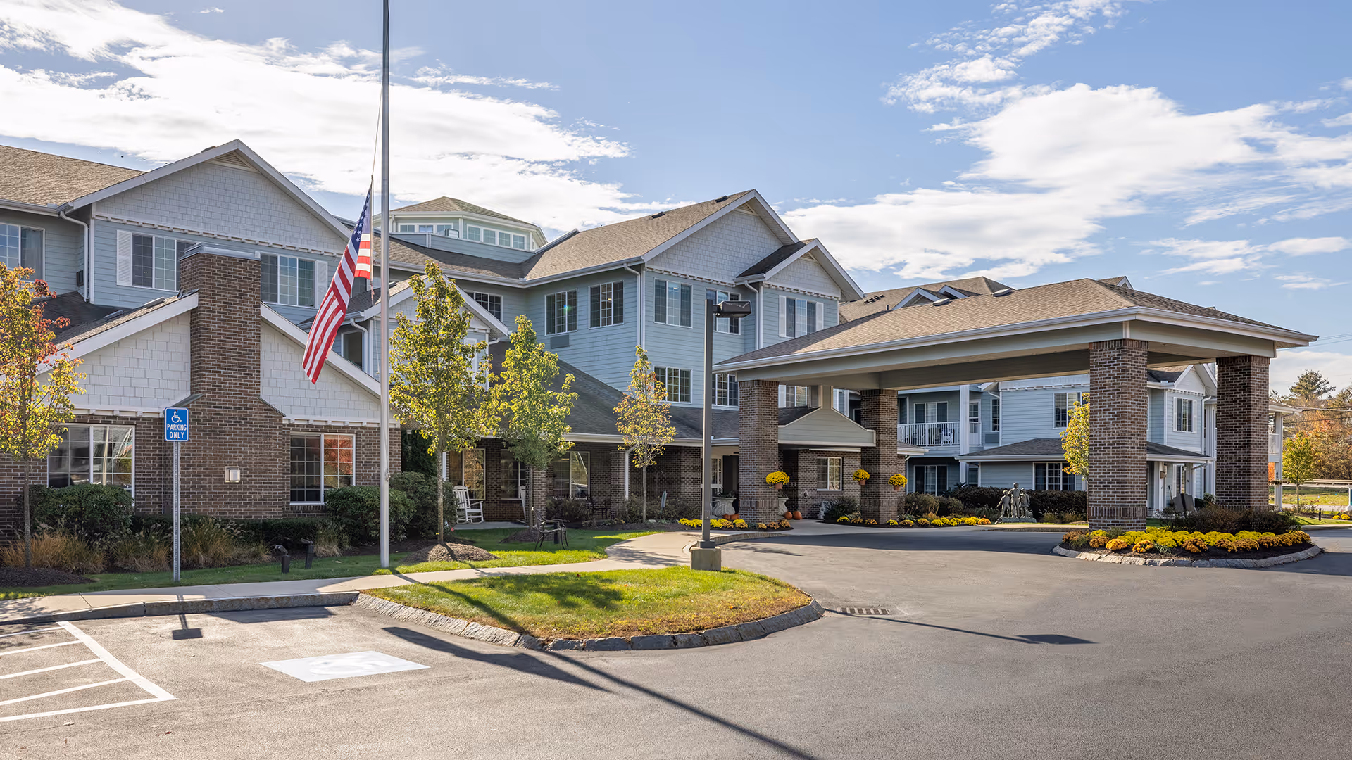 Exterior view of Holiday Kittery Estates senior living facility showing a large covered entrance with brick pillars, a flagpole with an American flag at half-mast, landscaped greenery, and a clear blue sky with some clouds.