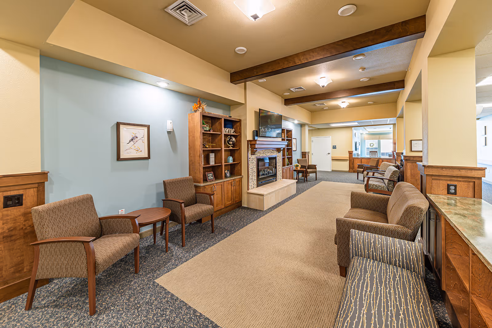 A cozy and well-lit common area in a senior living facility featuring multiple upholstered chairs and small tables arranged along the walls. There is a fireplace with a mounted TV above it, wooden shelves with decorative items, and a mix of carpet and wood paneling on the walls. The ceiling has exposed wooden beams and recessed lighting.