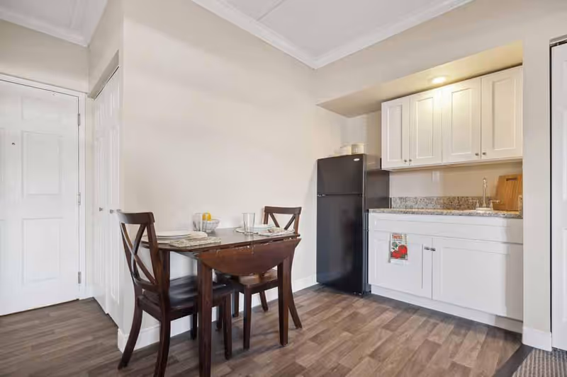 Small kitchen area with white cabinets, a black refrigerator, a granite countertop with a sink, and a wooden dining table set with two chairs and place settings. The floor is wood-patterned, and there is a white door and closet on the left side.