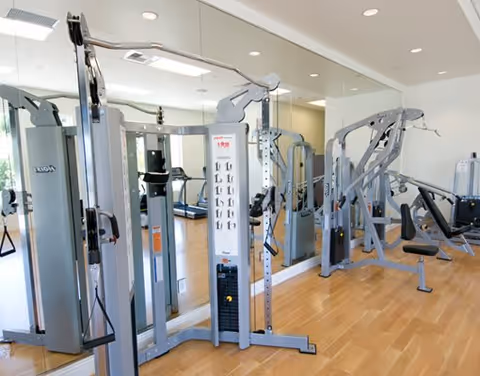 A bright and clean fitness room with various exercise machines including weight training equipment and cardio machines, with a large mirror covering one wall and wooden flooring.