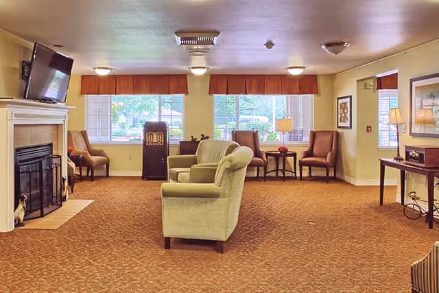 A cozy living room area with beige walls and carpeted floor. The room features a fireplace with a mounted TV above it on the left side, several armchairs arranged around the room, large windows with orange valances letting in natural light, a small table with a lamp, and framed artwork on the walls.