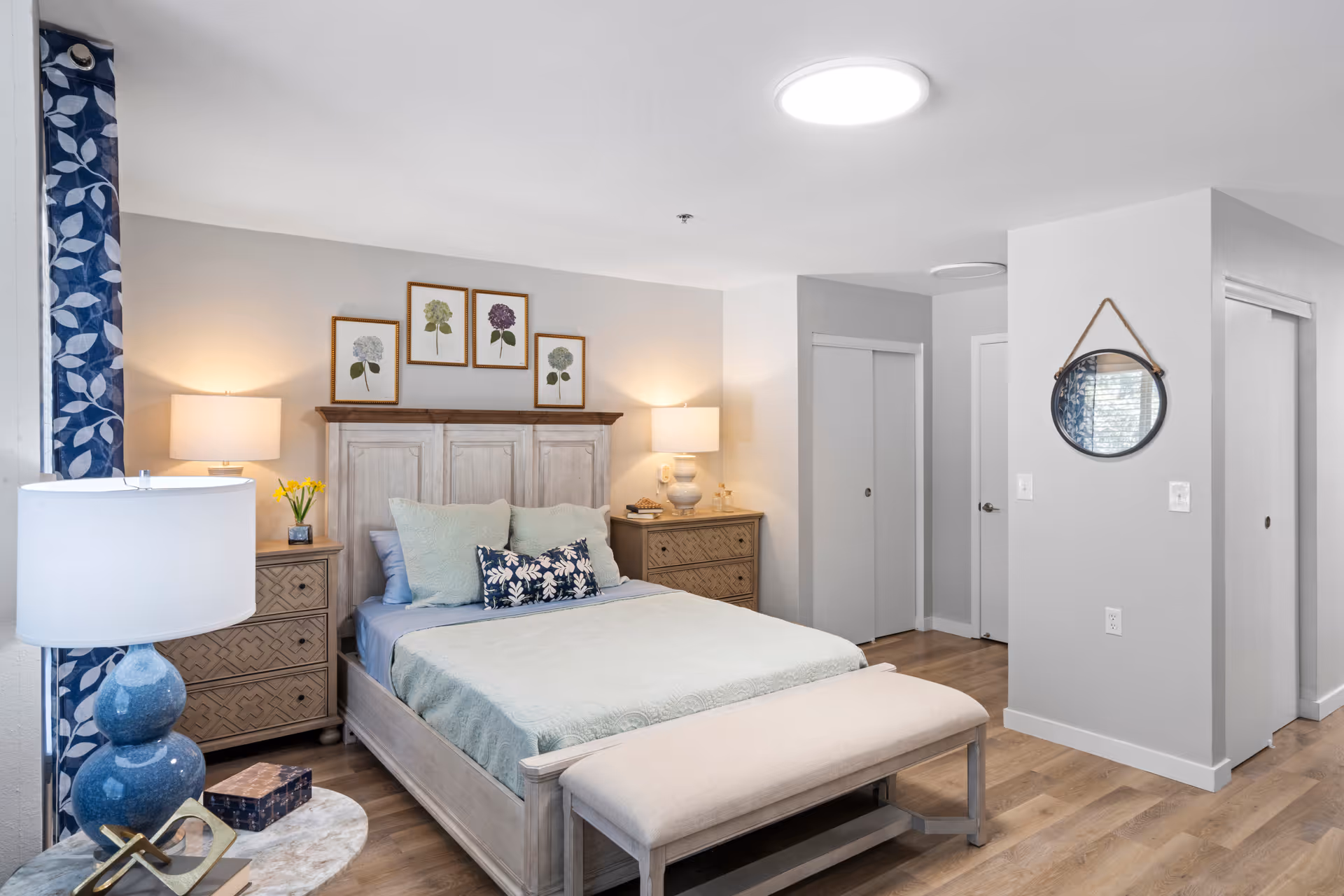A bright, furnished bedroom featuring a wooden bed with pillows and a bench, matching nightstands and lamps, framed botanical artwork above the headboard, and a round wall mirror.