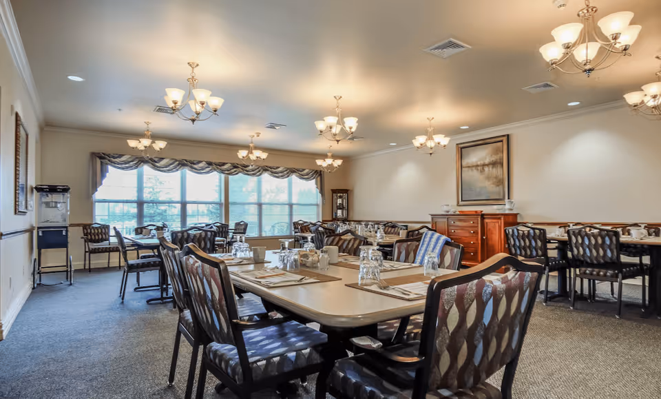 A well-lit dining room in a retirement village with multiple tables and chairs arranged neatly. Each table is set with placemats, glasses, and cups. Large windows with decorative valances allow natural light to fill the room. The walls are adorned with framed artwork and there are several chandeliers hanging from the ceiling providing warm lighting.