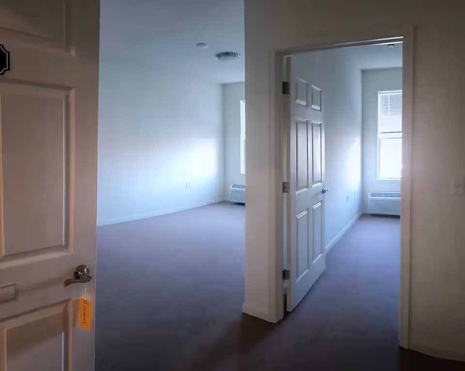 View of an empty apartment interior with beige carpet and white walls. The image shows an open door leading to a room with a window and a wall-mounted air conditioning unit below it. Another open door leads to a smaller room with a similar window and air conditioning unit.