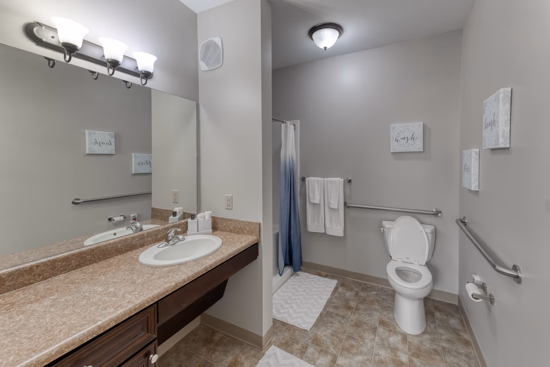 A clean and accessible bathroom with a single sink vanity featuring a large mirror and three-light fixture above. The bathroom has a toilet with grab bars on both sides, a shower with a blue and white curtain, and several white towels hanging on a towel rack. The walls are decorated with three small framed artworks with inspirational words.
