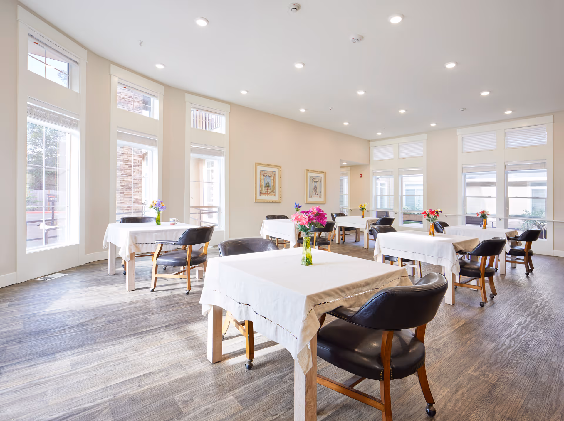 Bright dining room with multiple tables covered in white tablecloths, each adorned with a small vase of colorful flowers. The room has large windows allowing natural light to fill the space, wooden flooring, and several black cushioned chairs around the tables. The walls are light-colored with framed artwork hanging on one side.