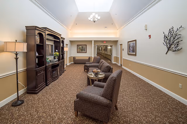 A well-lit interior common area with a skylight, featuring a dark wooden entertainment center with a TV, a floor lamp, two armchairs, a small round table with decorative items, and a sofa. The walls are painted beige with white trim, and there are framed pictures and a metal wall decoration. A hallway extends from the back of the room.