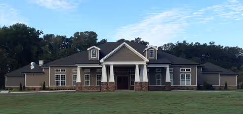 Front exterior view of a single-story building with a dark roof, beige siding, white columns, and multiple windows, set against a backdrop of trees and a partly cloudy sky.