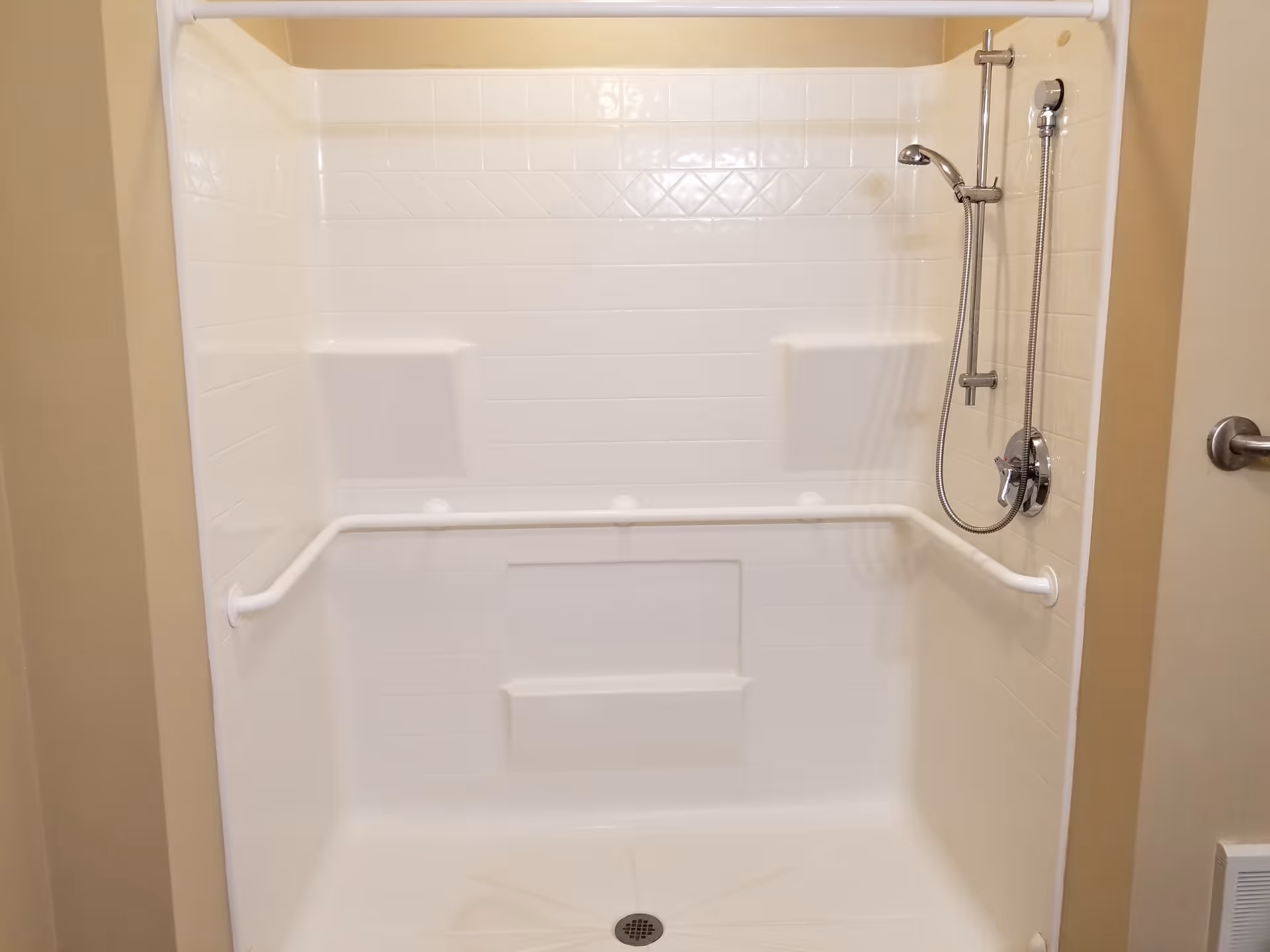 A clean, white shower stall with built-in shelves and a horizontal grab bar. The shower has a handheld showerhead attached to a vertical sliding bar, and the walls are tiled with a simple pattern.