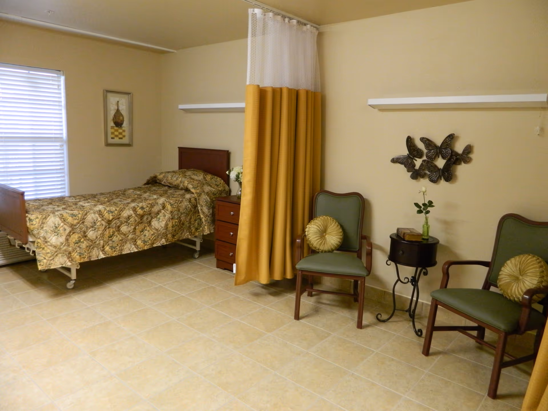 A nursing facility room with a single bed covered in a patterned bedspread, a wooden nightstand with a vase of flowers, two green cushioned chairs with round decorative pillows, a small round table with a flower vase and books, a yellow and white privacy curtain, and wall decorations including a framed picture and metal butterfly art.