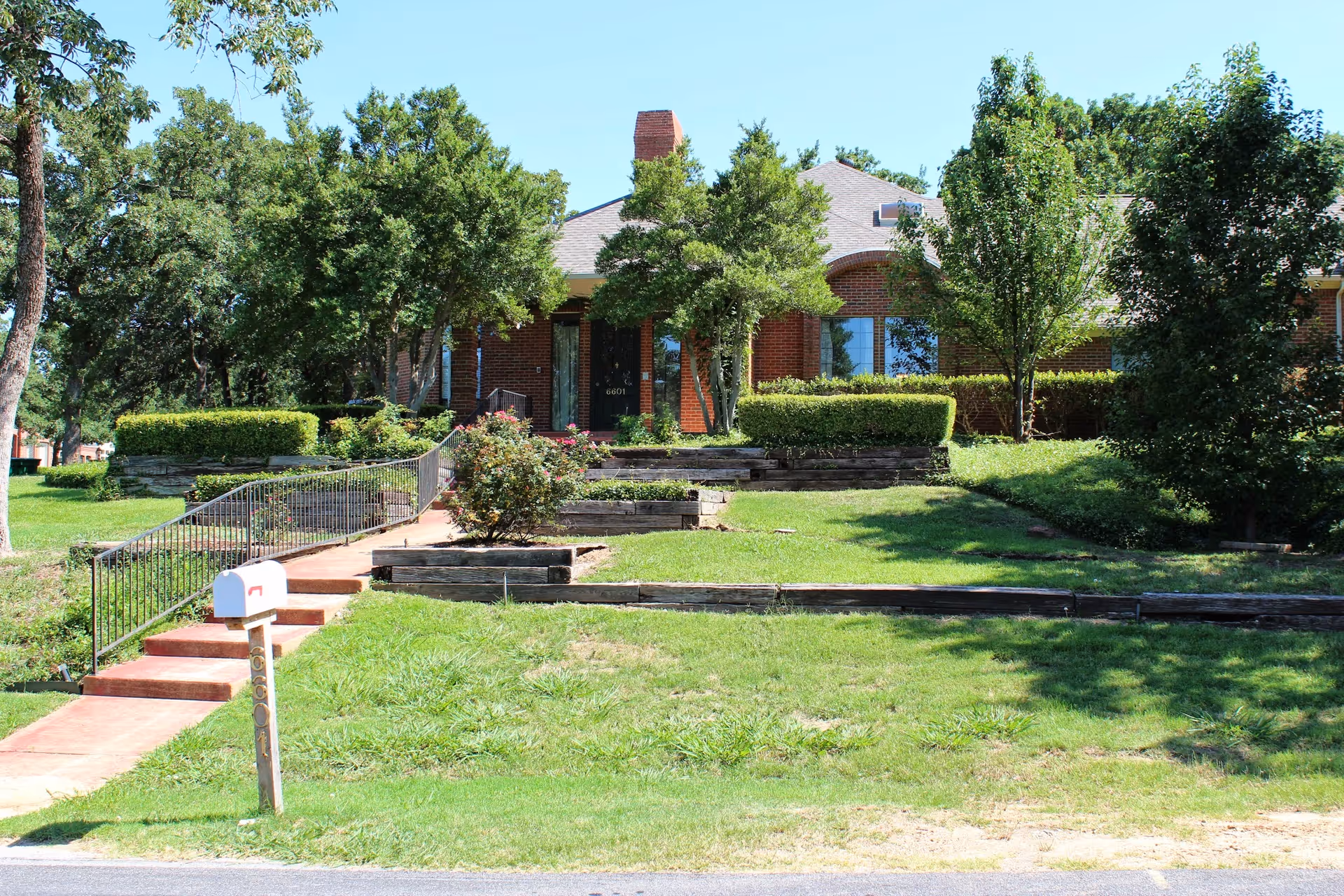 Front exterior view of a brick building partially obscured by trees and bushes, with a concrete walkway and steps leading up to the entrance. There is a white mailbox on a post near the street, and the lawn is green and well-maintained under a clear blue sky.