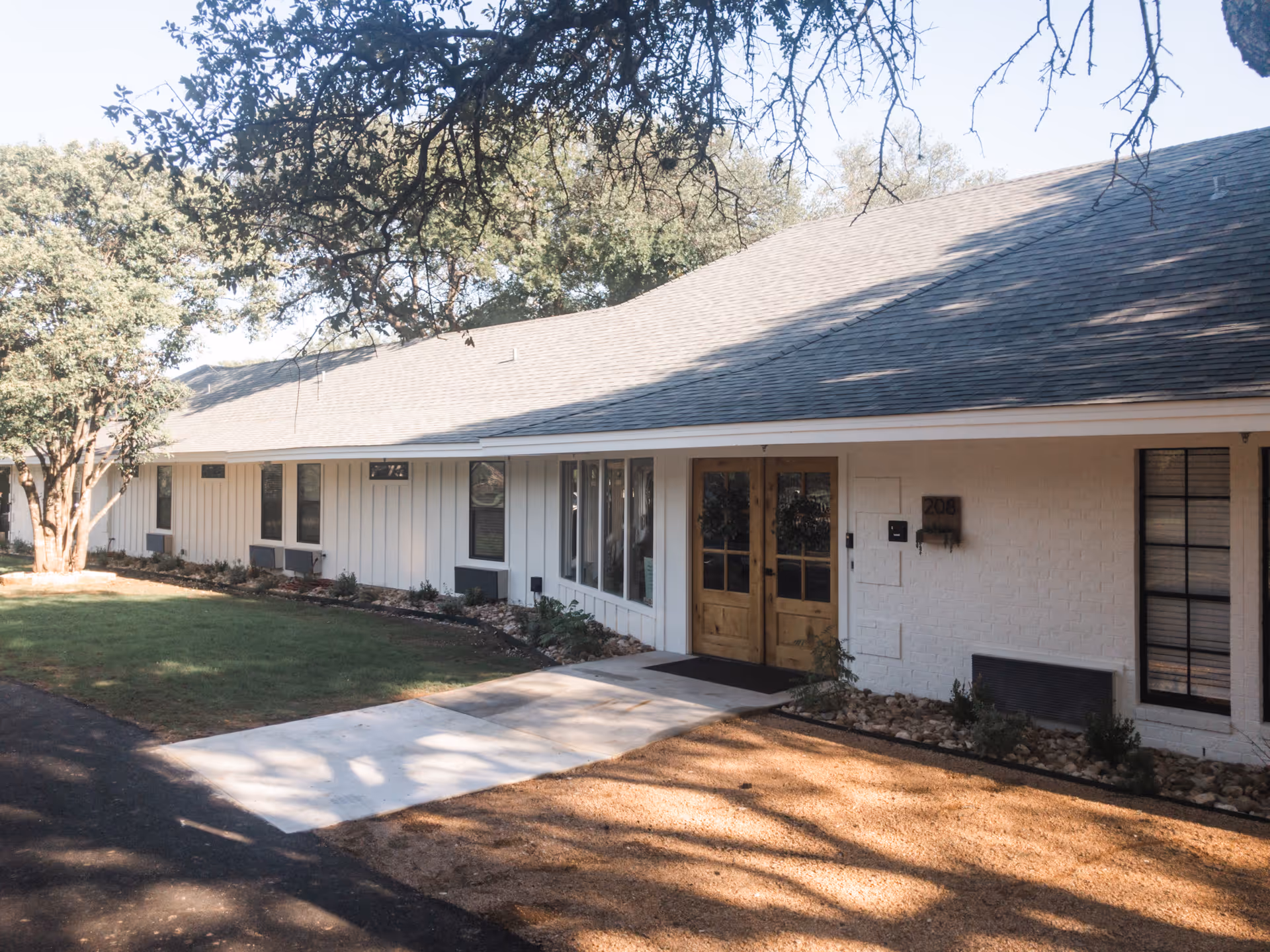 Exterior view of a single-story building with white siding and a gray shingled roof, featuring a wooden double door entrance with the number 208 on the wall next to it. There are several windows along the building and a small landscaped area with grass, rocks, and trees casting shadows on the ground.