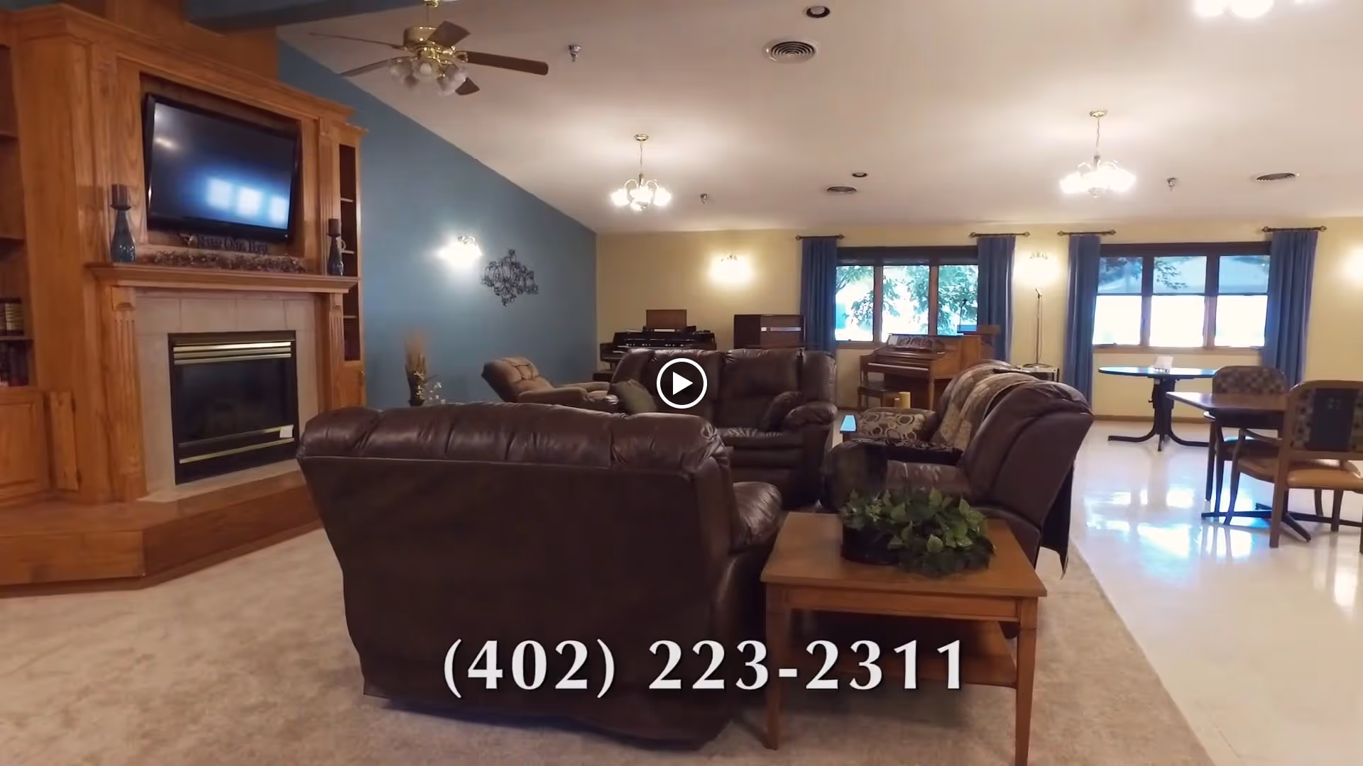 A spacious living room area with brown leather sofas and armchairs arranged around a wooden coffee table with a plant. There is a large wooden fireplace with a mounted flat-screen TV above it on the left side. The room has blue and beige walls, ceiling fans, and multiple light fixtures. In the background, there are large windows with blue curtains, a piano, and dining tables with chairs.