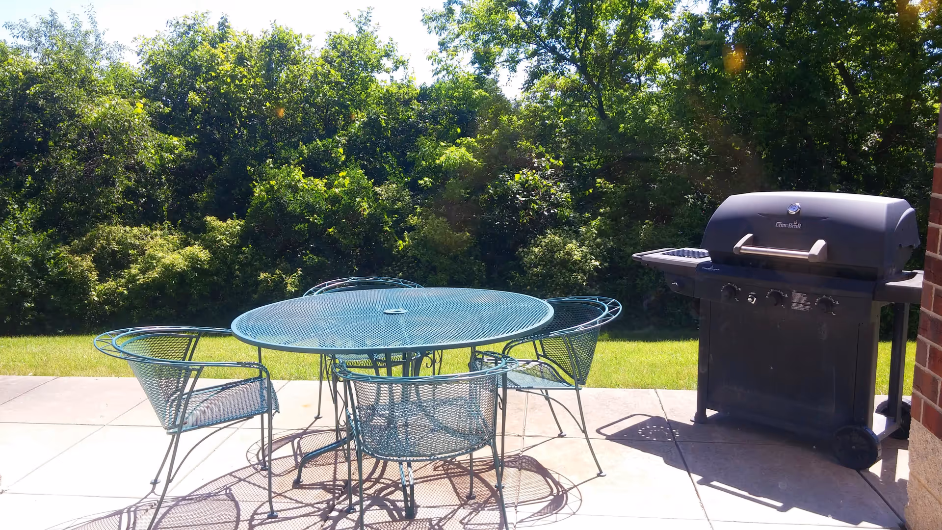 Outdoor patio area with a round metal table and four matching metal chairs on a tiled surface, next to a black gas grill. In the background, there is green grass and dense trees under a sunny sky.