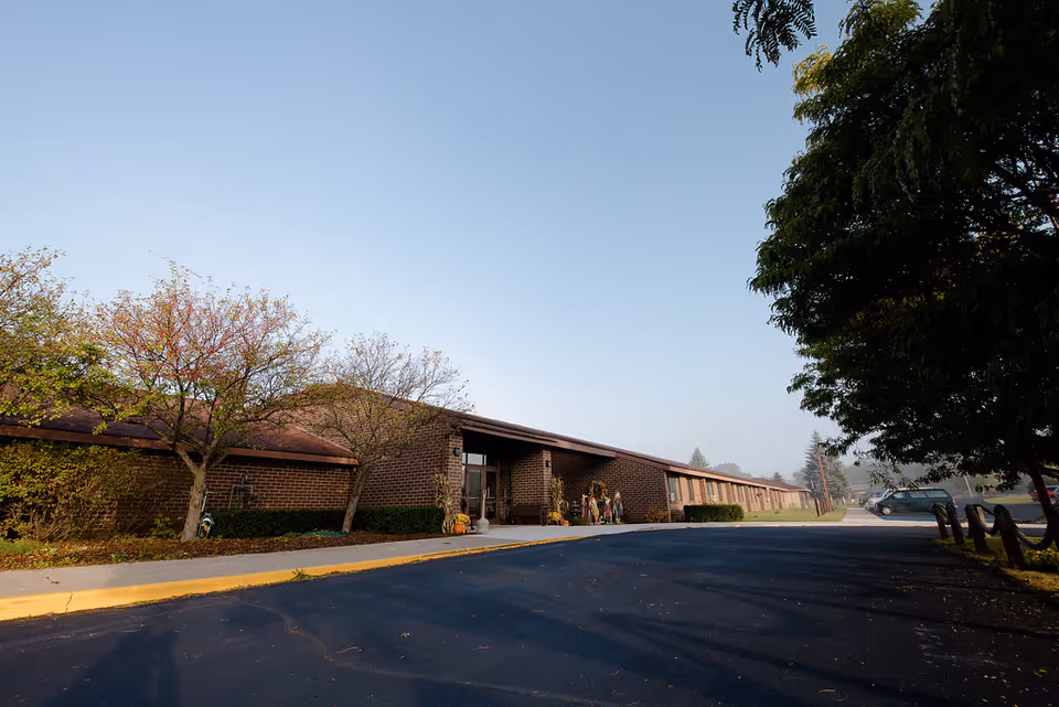 Exterior view of a single-story brick building with a sloped roof, surrounded by trees and a paved driveway. The entrance is decorated with autumn-themed items including pumpkins and cornstalks. A few cars are parked in the distance under a clear blue sky.