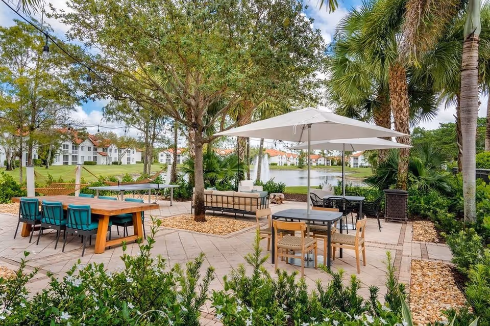 Outdoor patio area with tables, chairs, umbrellas and lounge seating by a pond and fountain, surrounded by trees and nearby residential buildings.