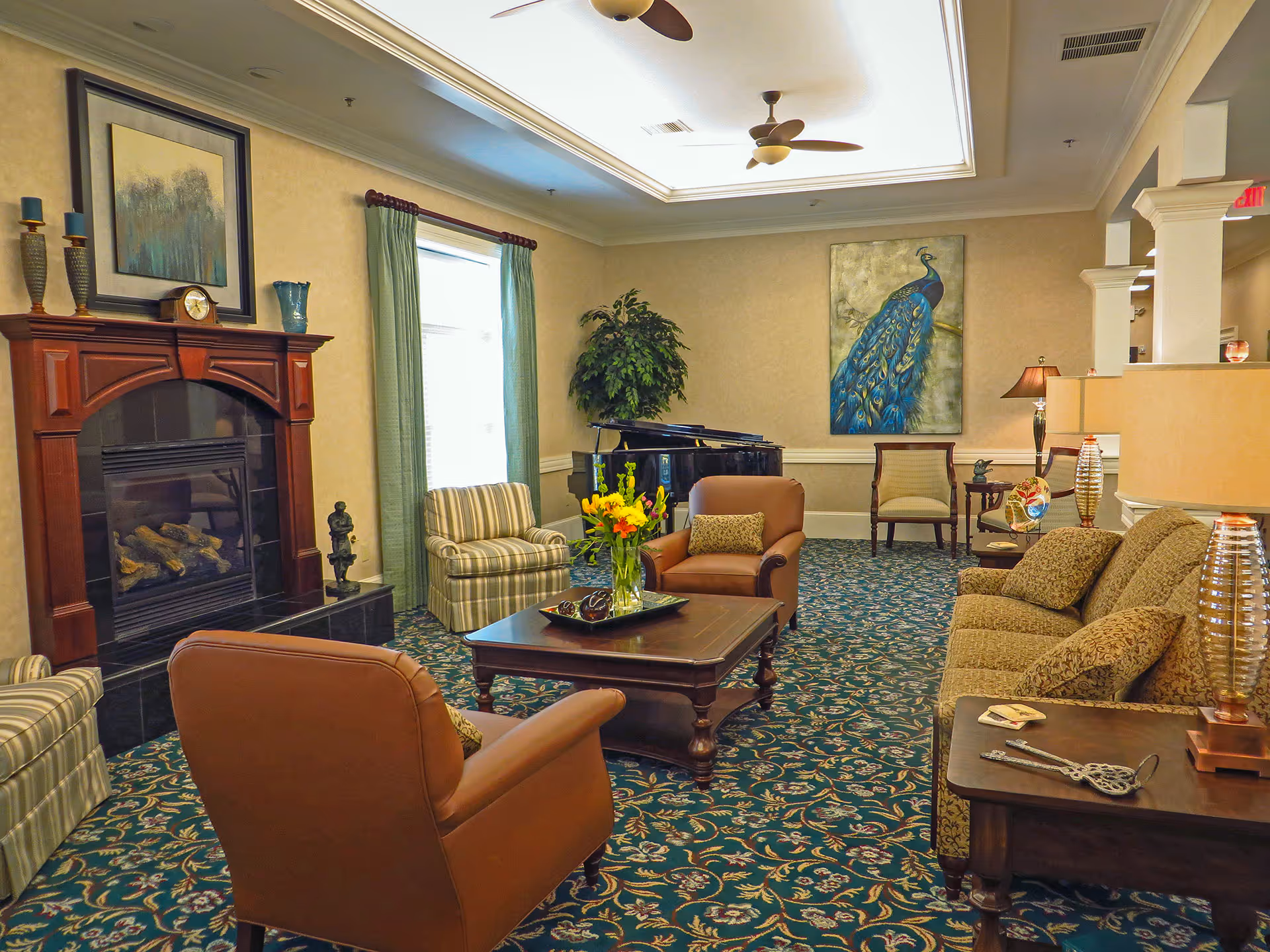 A cozy living room with patterned carpet, a wooden coffee table with a flower vase, two brown armchairs, a patterned sofa, and a fireplace with decorative items on the mantel. There is a large painting of a peacock on the wall, a grand piano in the corner, and ceiling fans above.