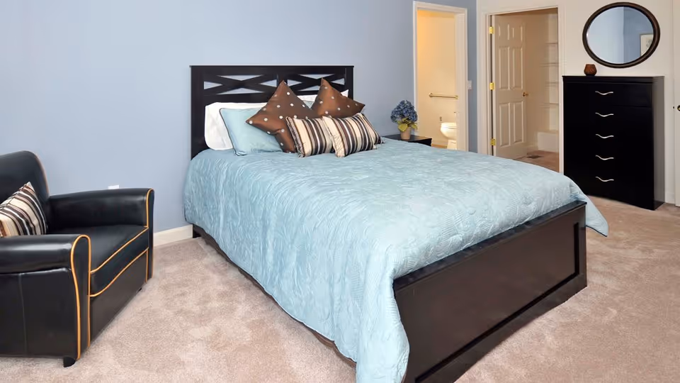 A bedroom with a large bed covered in a light blue quilt and several decorative pillows. To the left of the bed is a black armchair with a striped pillow. In the background, there is a doorway leading to a bathroom with a visible toilet. On the right side of the room, there is a black dresser with a round mirror above it and a small decorative item on top. The walls are painted light blue and the floor is carpeted.