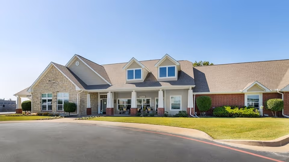 Exterior view of a single-story senior living facility building with a combination of stone and brick facade, a covered entrance with columns, and neatly trimmed bushes and lawn under a clear blue sky.