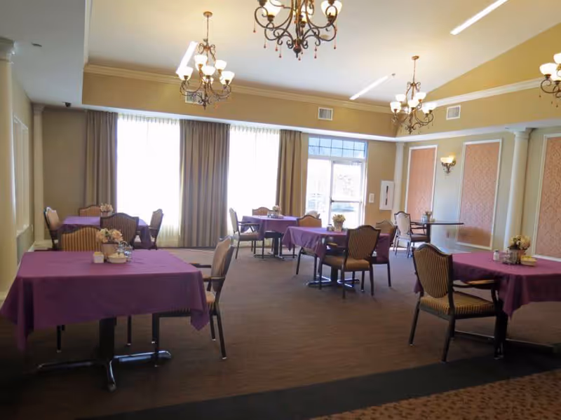 A dining room with several tables covered in purple tablecloths, each surrounded by chairs. The room has large windows with curtains allowing natural light to enter, chandeliers hanging from the ceiling, and decorative wall panels with sconces.