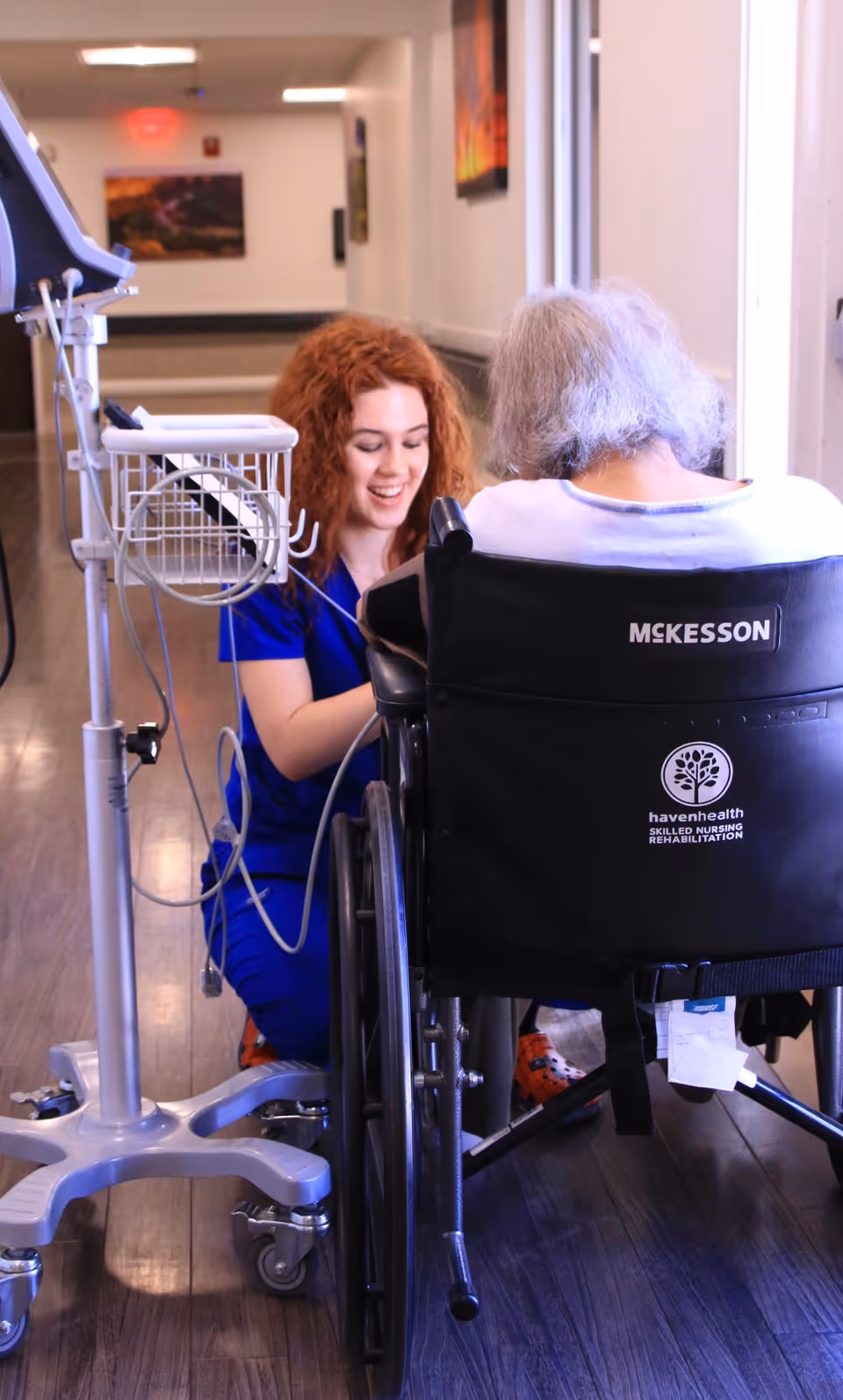 A healthcare worker in blue scrubs kneels beside an elderly person seated in a wheelchair in a hallway. The healthcare worker is smiling and appears to be interacting warmly with the elderly person. Medical equipment is visible next to them. The wheelchair has the logos 'McKesson' and 'havenhealth Skilled Nursing Rehabilitation' on the back.