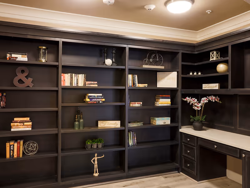 Interior view of a room with dark wooden built-in shelves and cabinets. The shelves contain books, decorative items such as a small bicycle sculpture, candles, plants, and signs with words like 'Family' and 'Laugh'. There is a white countertop with drawers underneath and a potted orchid plant on the right side. The ceiling has recessed lighting and crown molding.