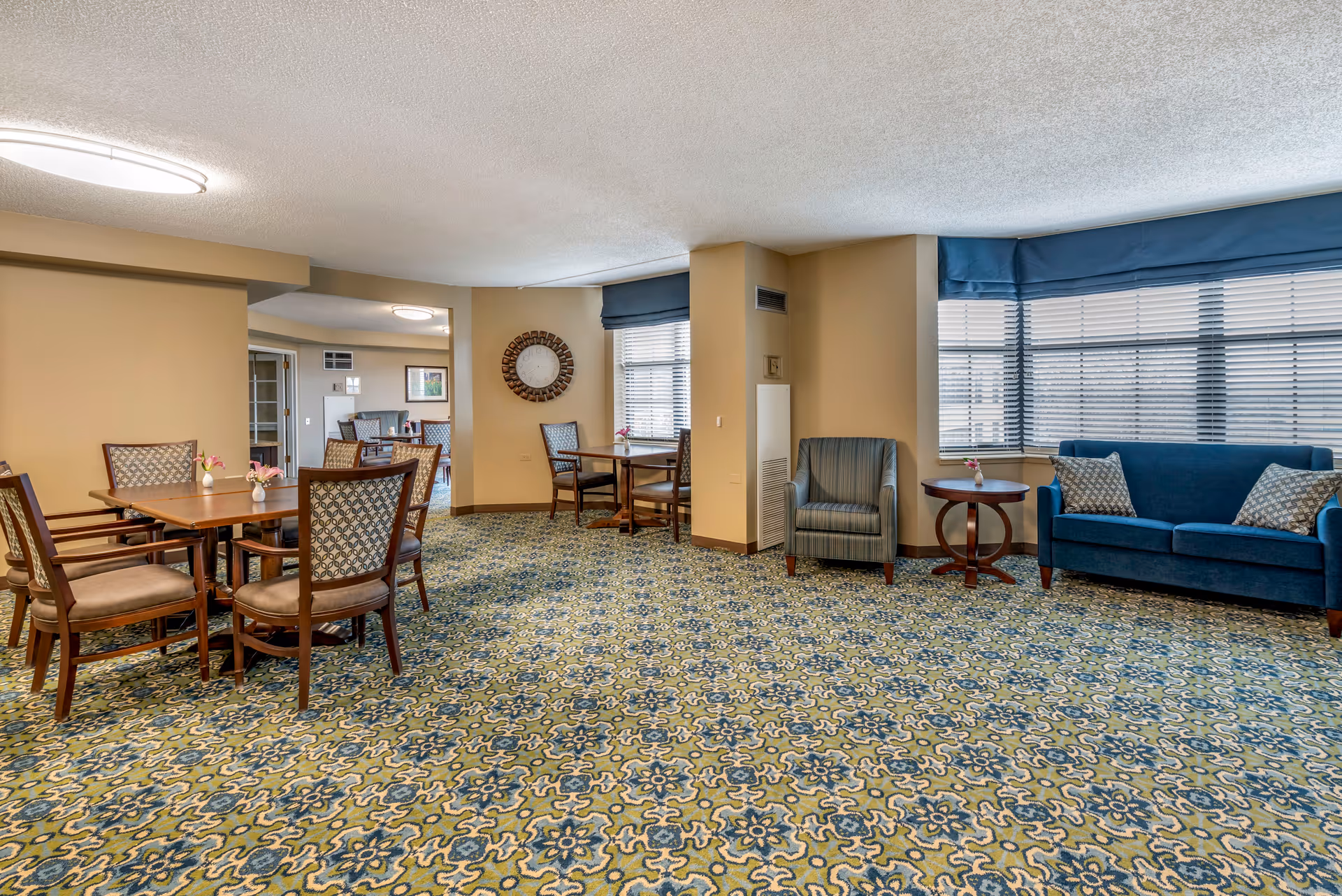 A spacious common area in a senior living facility featuring patterned carpet, several wooden tables with chairs, a blue sofa with patterned cushions, a striped armchair, and large windows with blinds and blue valances. A round wall clock is mounted on a beige wall.