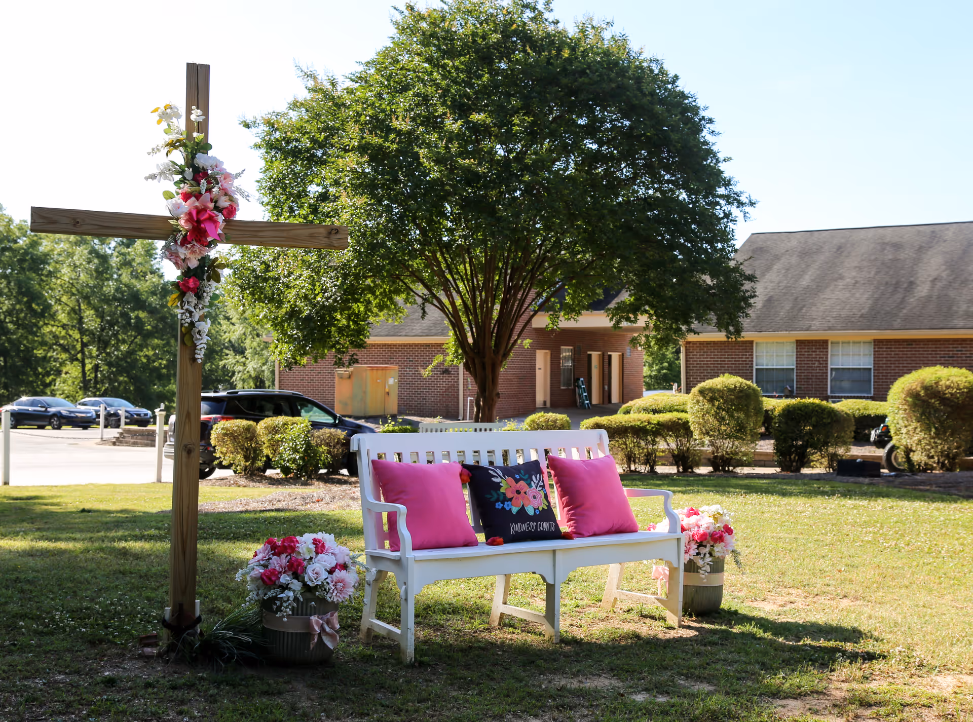 Outdoor scene at Green Acres Health & Rehabilitation featuring a white wooden bench with pink and floral cushions placed on grass. Next to the bench is a wooden cross decorated with flowers, and flower arrangements are placed on the ground nearby. In the background, there are trimmed bushes, a large tree, parked cars, and brick buildings under a clear blue sky.