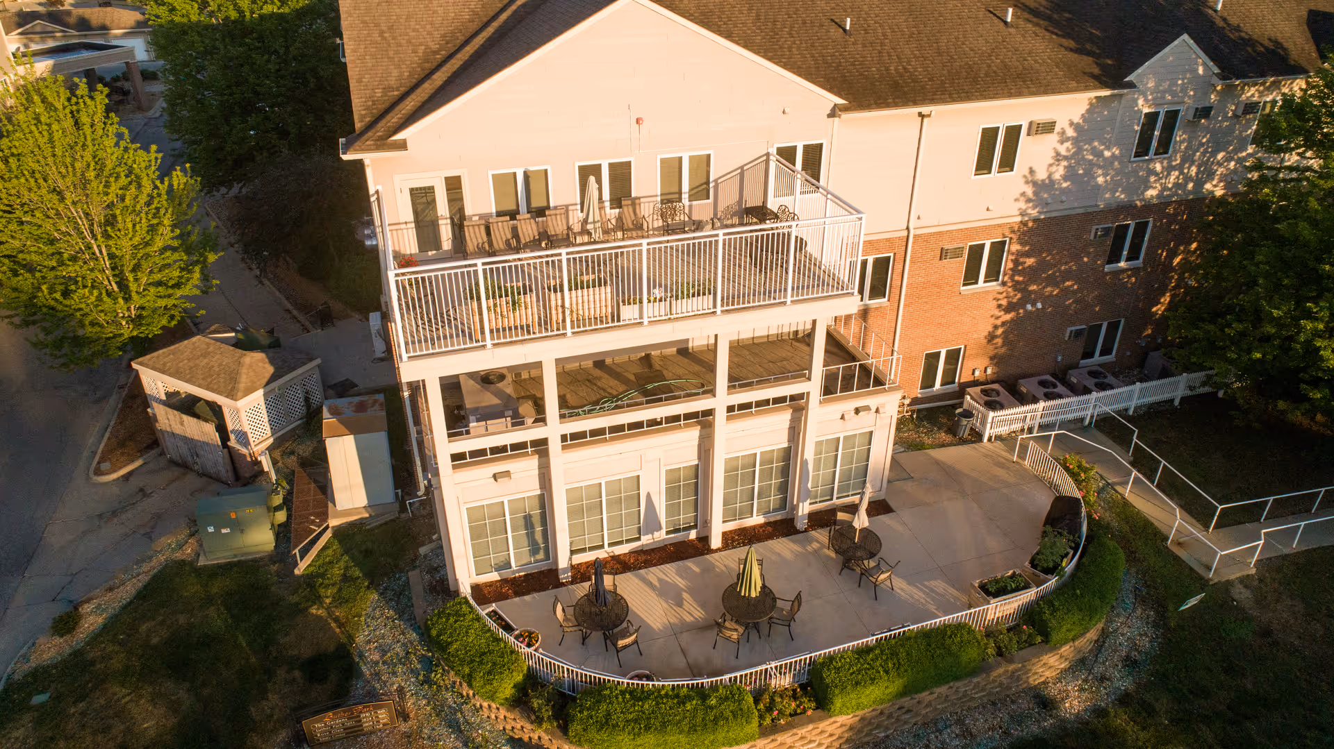Aerial view of a multi-story building with a large patio area featuring round tables and chairs with umbrellas. The building has a balcony on the second floor with additional seating. Surrounding the patio is a white fence and landscaped greenery. The building exterior is a combination of brick and light-colored siding.