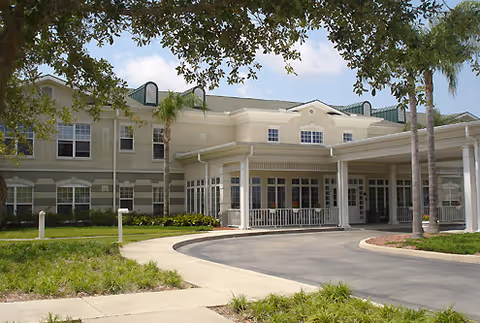 Front entrance of a two-story senior living facility with a covered porte-cochère, columns, palm trees, and landscaped grounds.