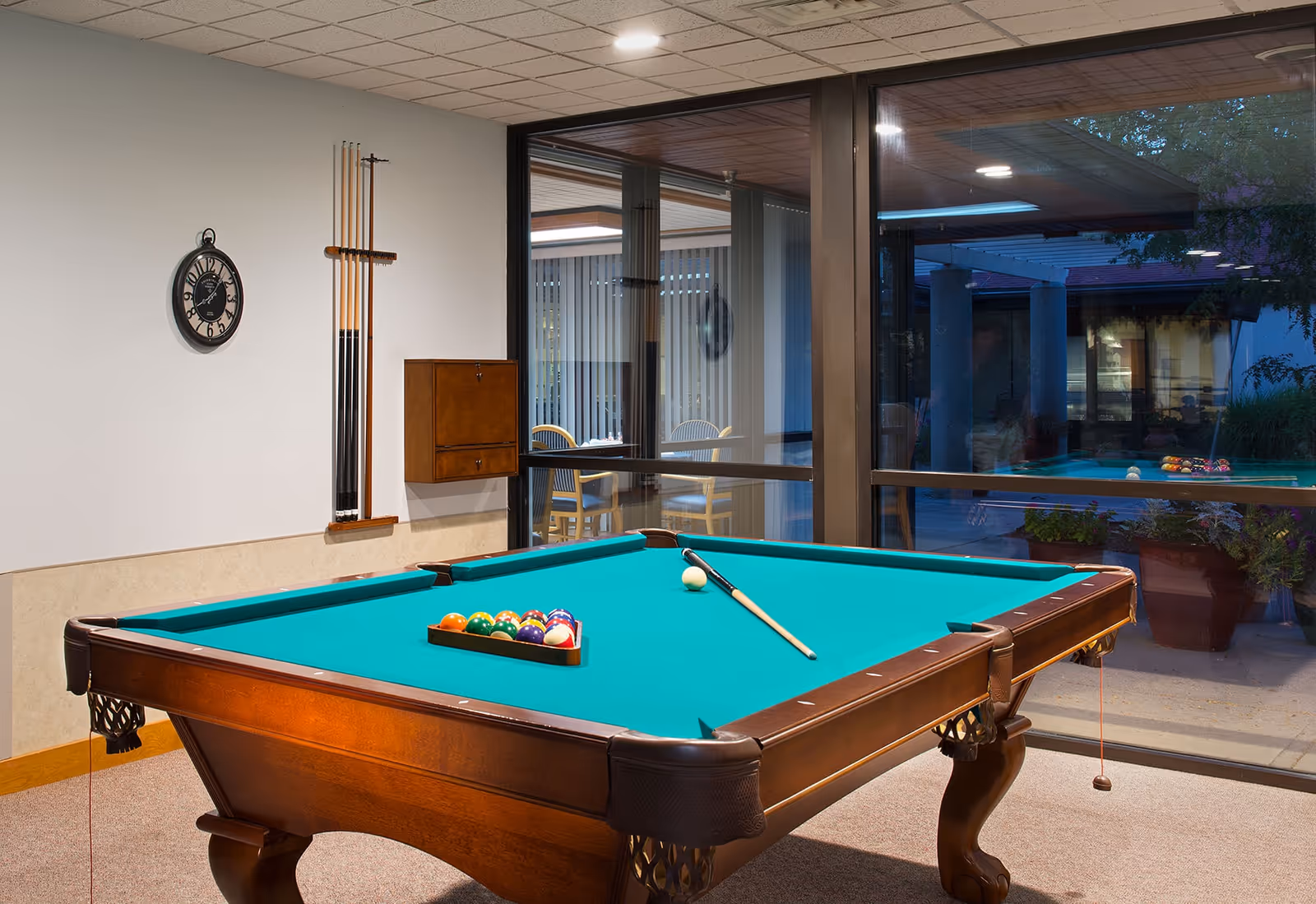 A billiards table with cues and a racked set of balls in a glass-walled recreation room.