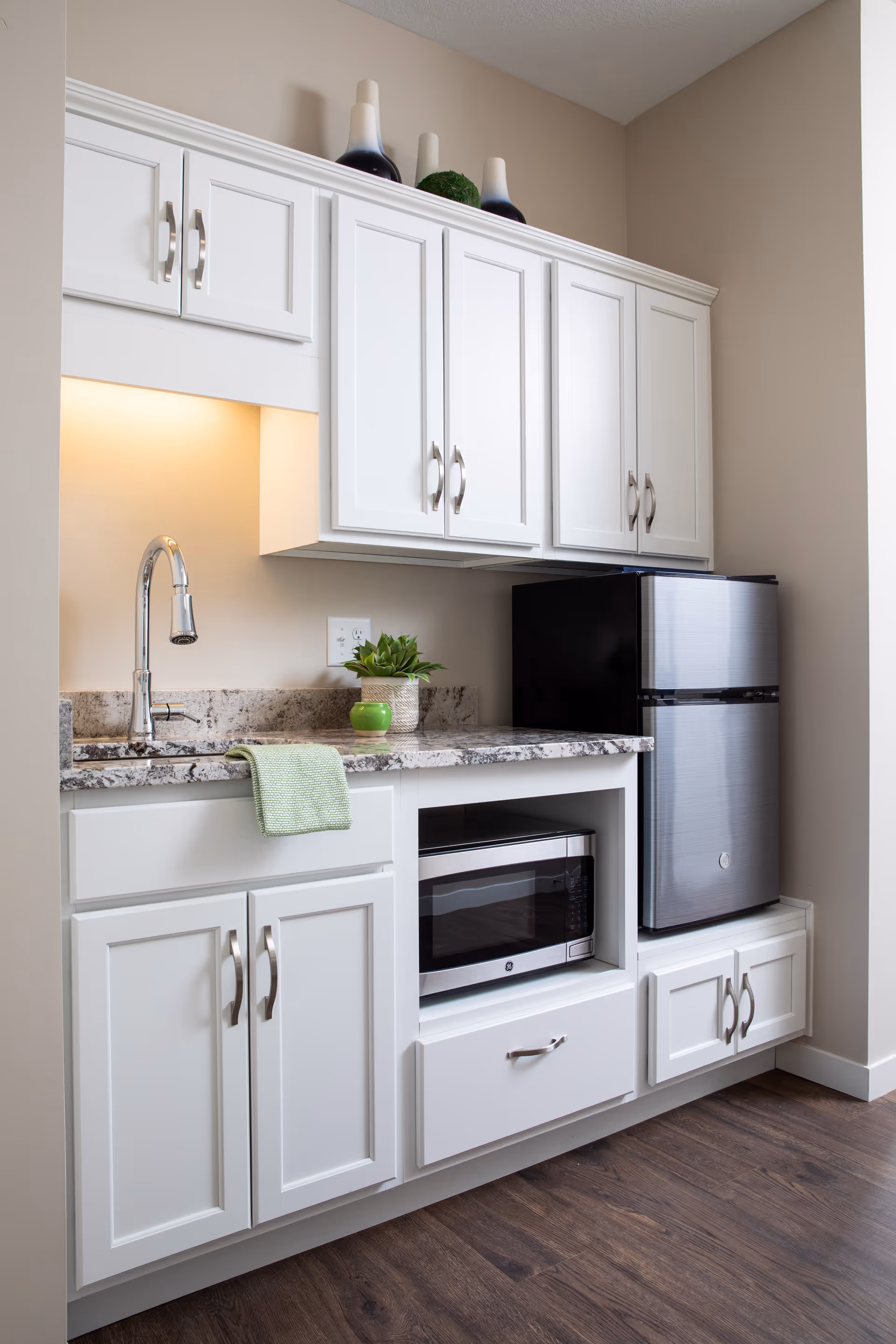 A small kitchen area with white cabinets, a granite countertop, a stainless steel sink with a chrome faucet, a microwave oven built into the cabinetry, and a compact stainless steel refrigerator. There are decorative items on top of the cabinets and a green towel hanging over the edge of the counter.