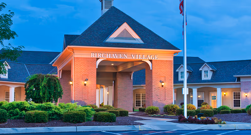 Exterior view of Birchaven Village building entrance at dusk, featuring brick walls, a covered driveway, well-maintained landscaping with bushes and small trees, and an American flag on a flagpole.