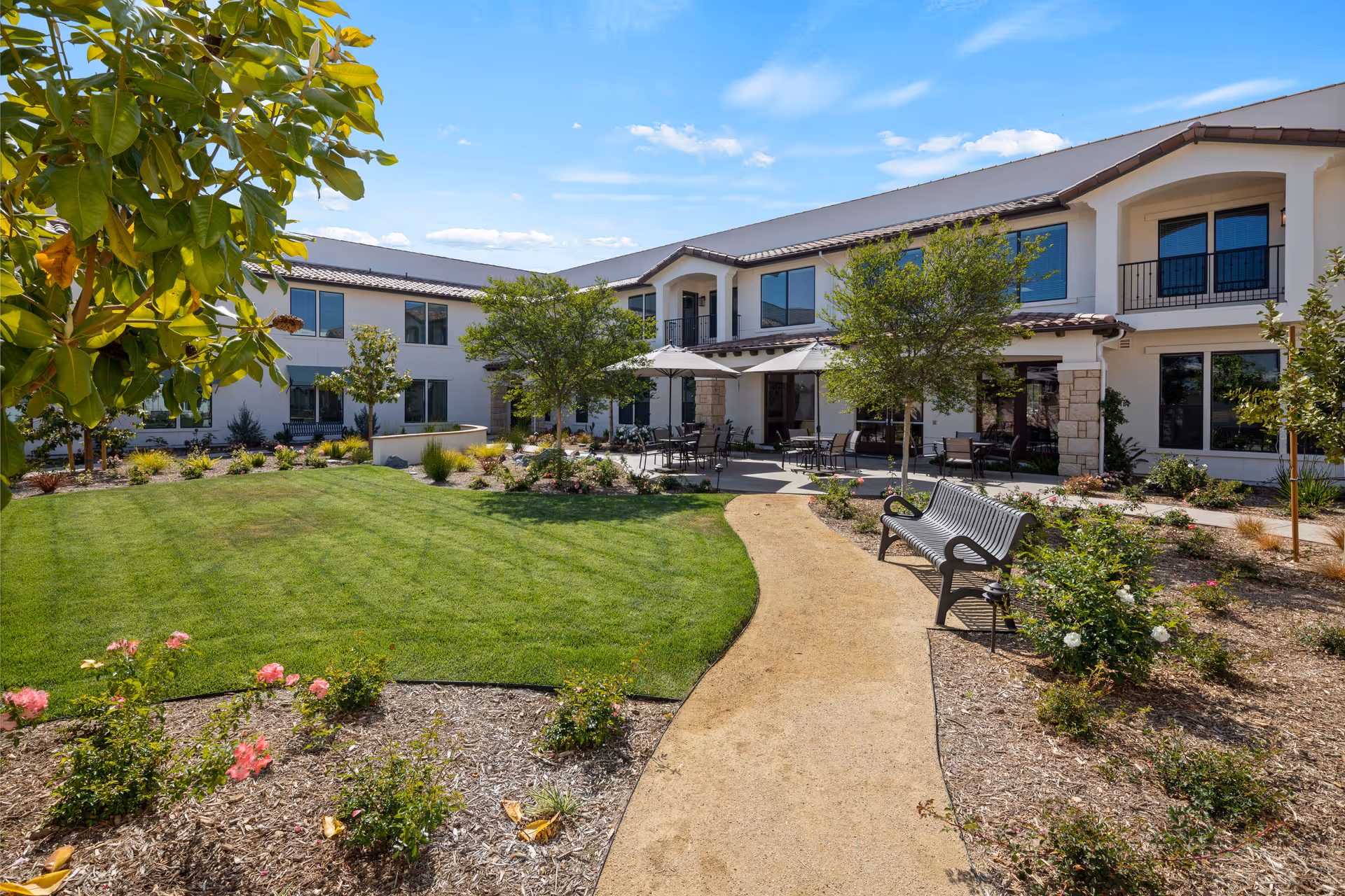 Outdoor courtyard area at Oakmont of Simi Valley featuring a curved walking path, green lawn, flower beds with blooming roses, several trees, benches, and patio tables with umbrellas in front of a two-story building with large windows.
