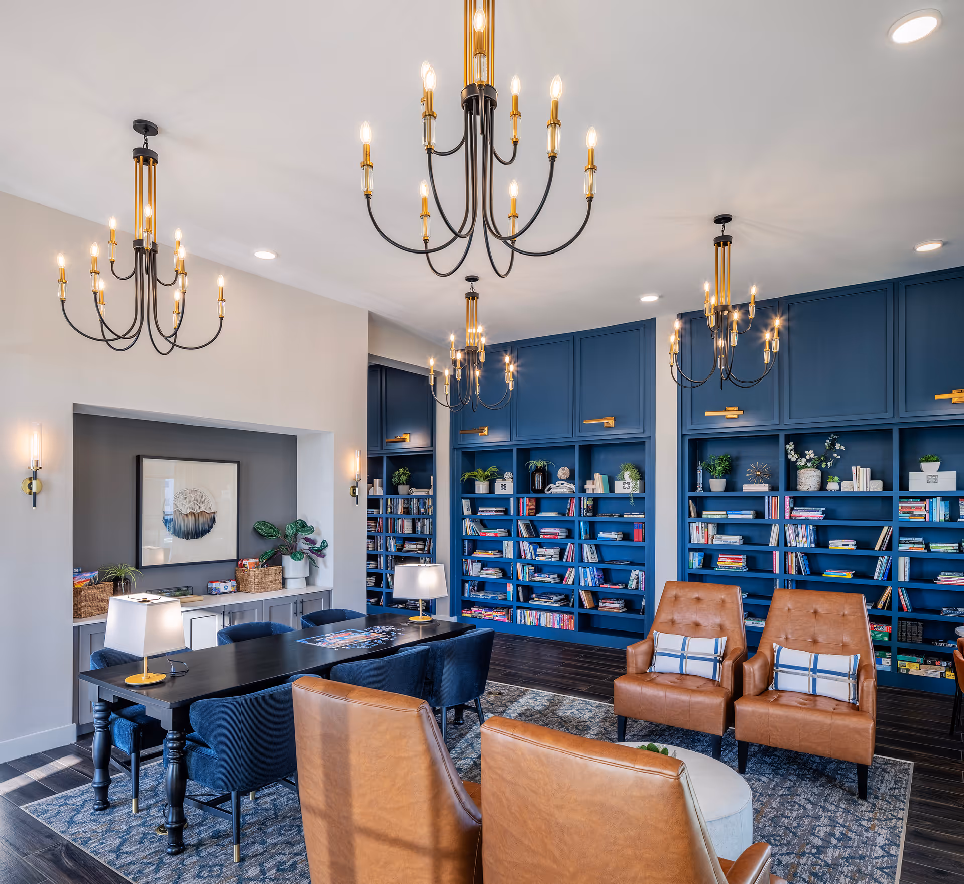 A stylish and cozy common area with blue built-in bookshelves filled with books and decorative items, a long black table with blue upholstered chairs, and four brown leather armchairs with plaid pillows arranged around a small round table. The room features multiple elegant chandeliers and wall sconces providing warm lighting, with a framed artwork and plants on a sideboard against a light-colored wall.