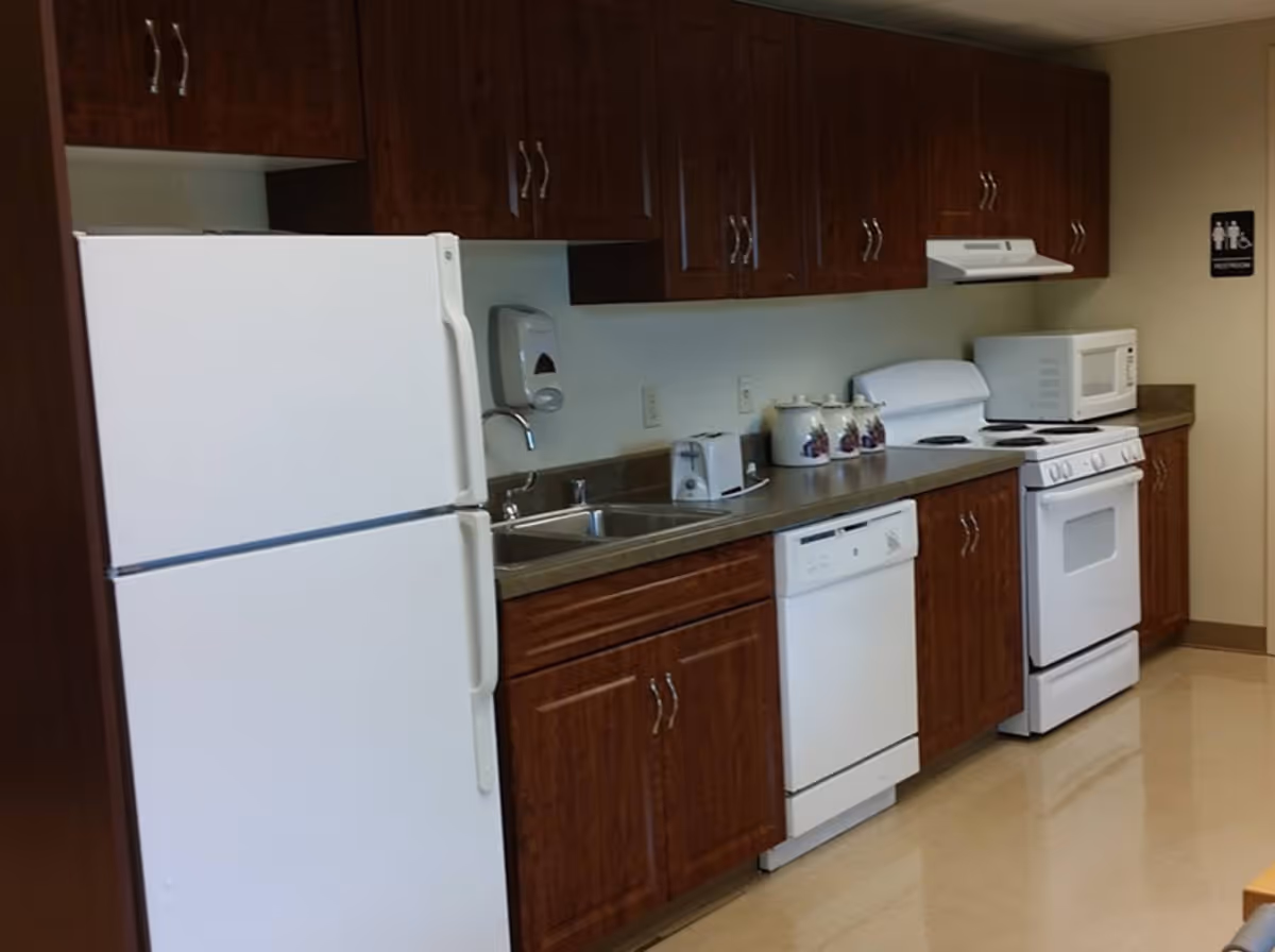 A kitchen area with white appliances including a refrigerator, dishwasher, stove, and microwave. The kitchen has dark wood cabinets above and below the countertop, a double sink, a toaster, and three decorative canisters on the counter. There is a restroom sign on the wall in the background.