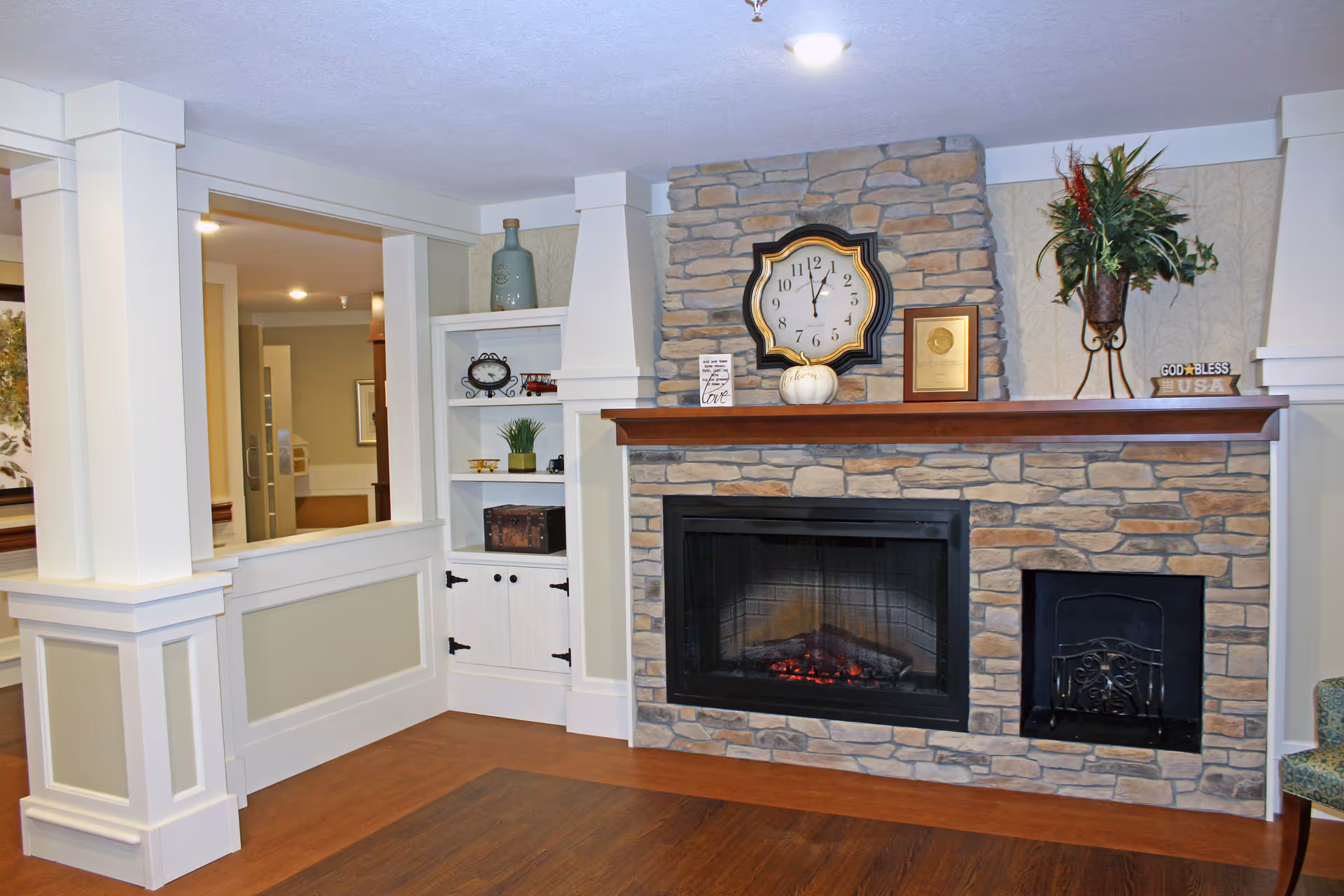 Stone fireplace with a wooden mantel decorated with a clock, plants, and decor in a cozy common room.