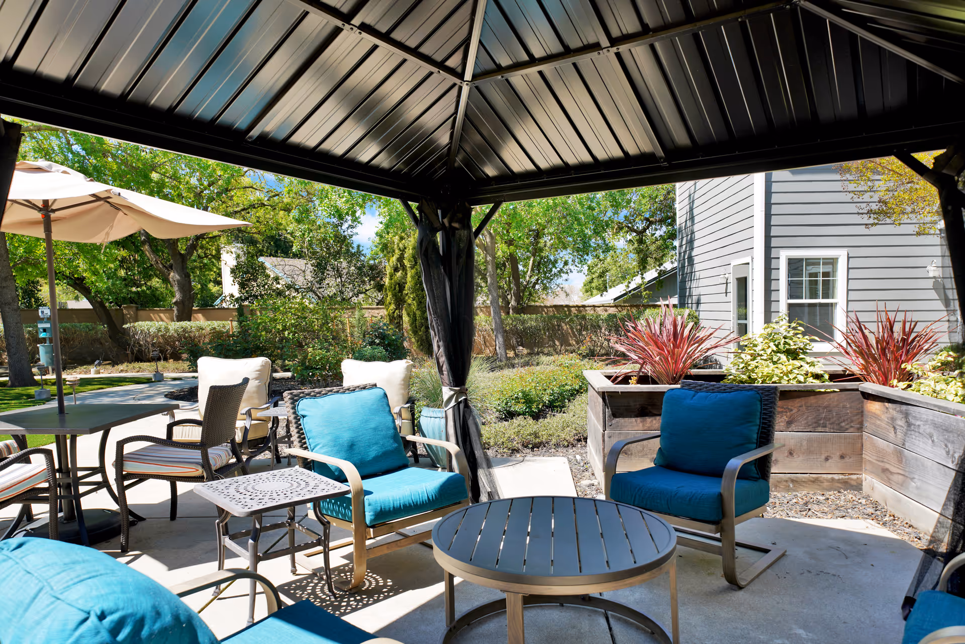 Outdoor seating area under a metal gazebo with blue cushioned chairs and a round table. Surrounding the area are plants, trees, and a building with gray siding and white trim. There is also a patio table with an umbrella and chairs in the background.