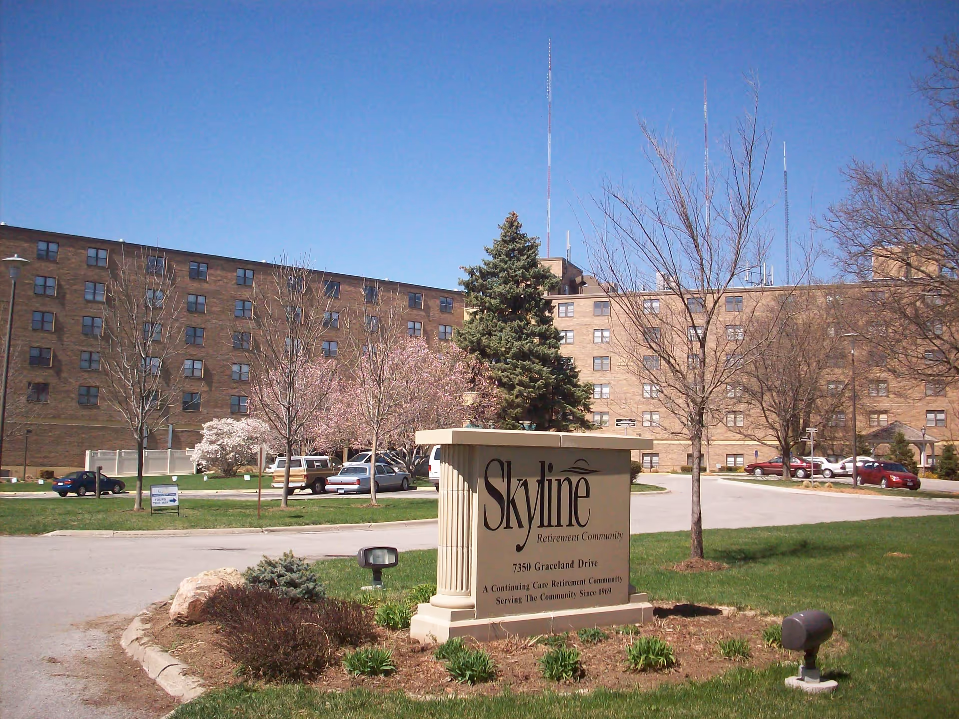 Exterior view of a large multi-story brick retirement community building with several windows. In the foreground, there is a landscaped area with a stone sign that reads 'Skyline Retirement Community, 7350 Graceland Drive, A Continuing Care Retirement Community Serving The Community Since 1969.' There are trees, some with blossoms, and several parked cars visible around the building under a clear blue sky.