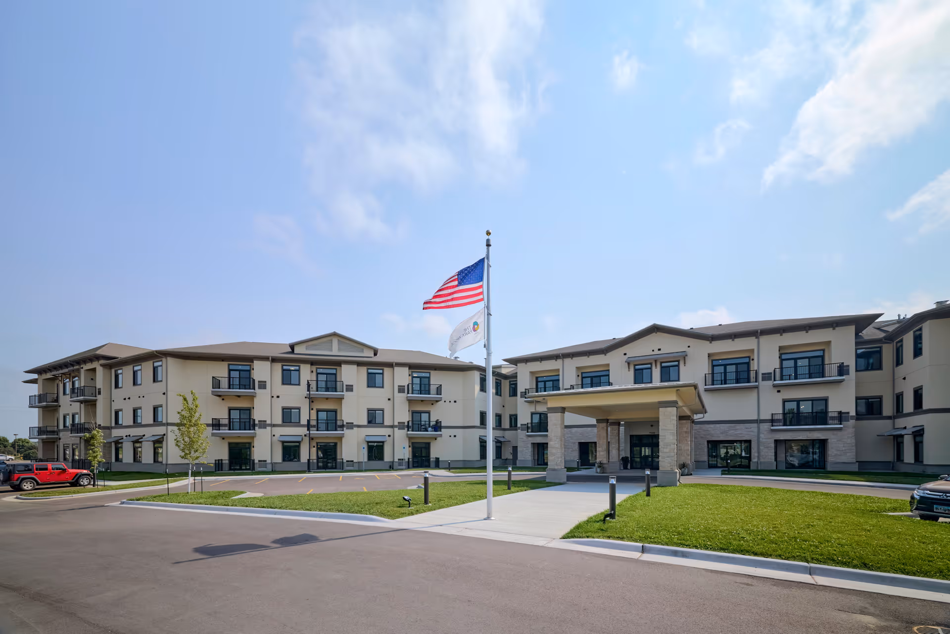Exterior view of The Parkwood senior living facility showing a modern three-story building with balconies, a covered entrance, a flagpole with the American flag and another flag, a parking area, and a well-maintained lawn under a partly cloudy sky.
