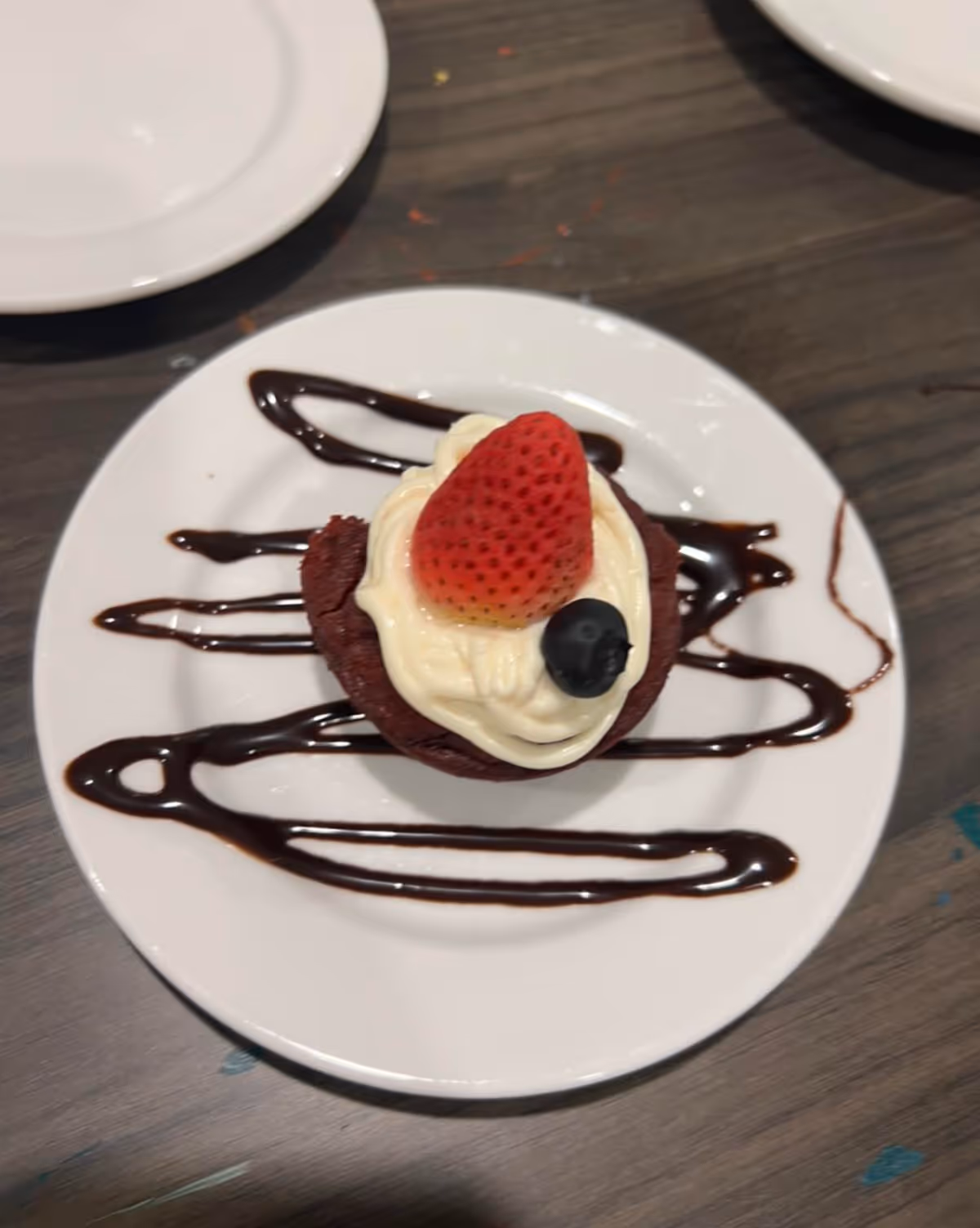 A small dessert topped with cream, a strawberry and a blueberry with chocolate drizzle on a white plate on a wooden table.