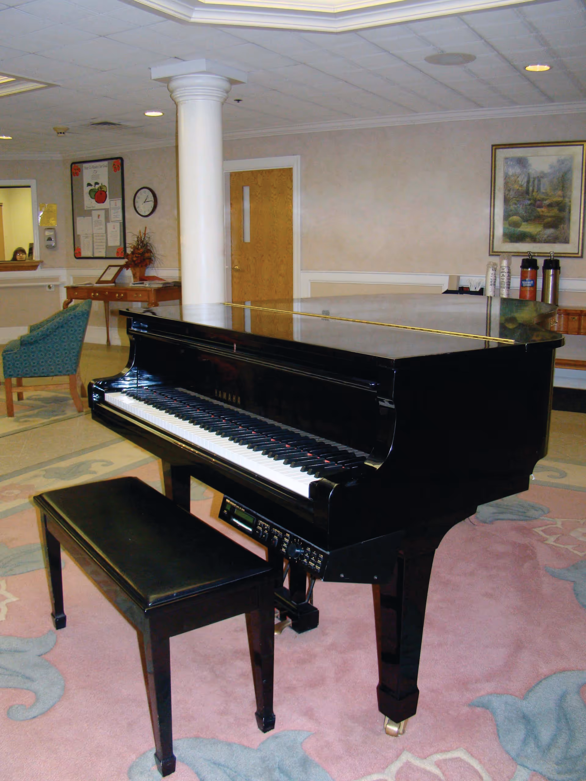 Black grand piano and bench in a senior facility lounge area with seating, column, and framed art.