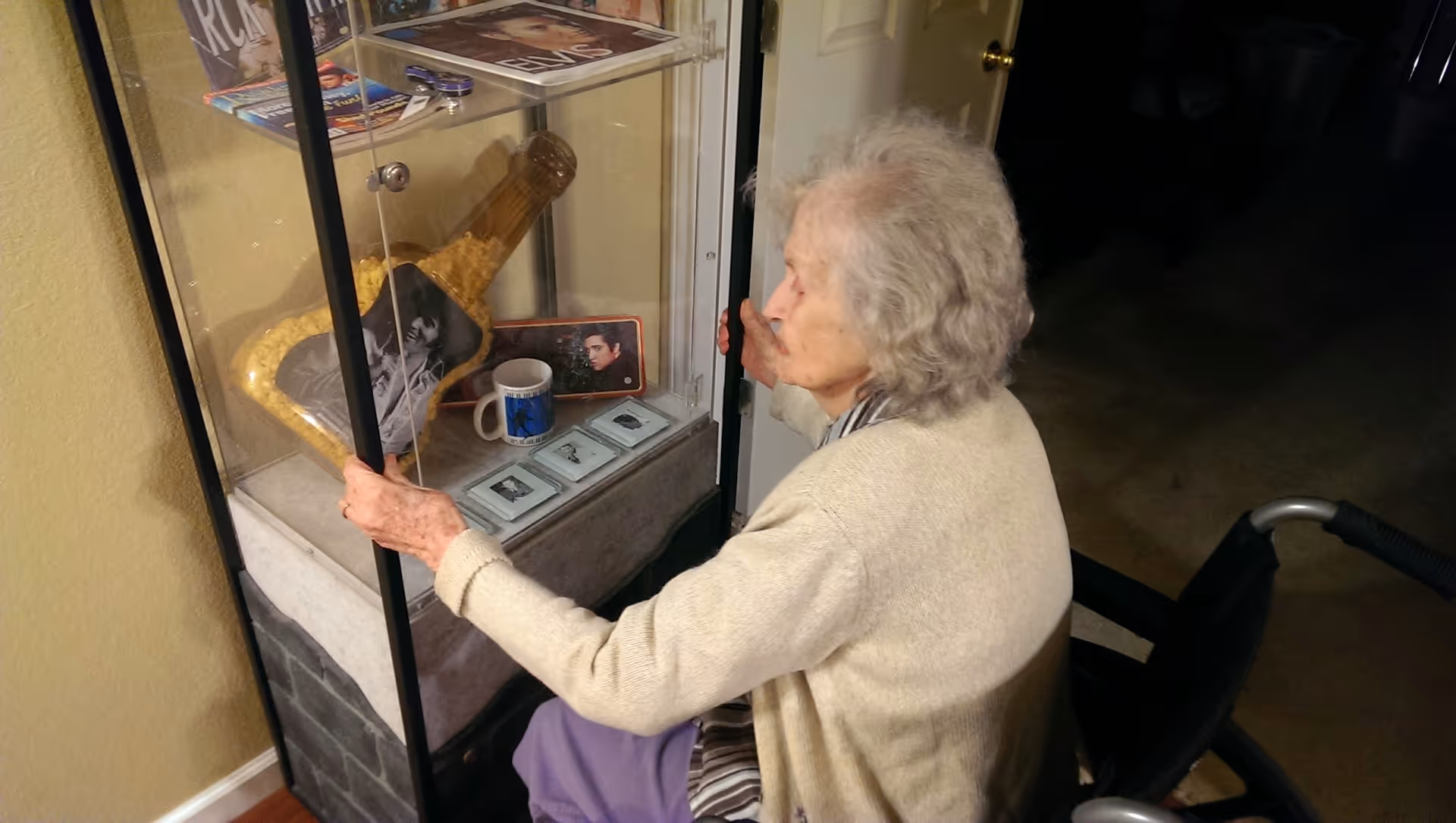 An elderly woman in a wheelchair is looking at and opening a glass display case containing memorabilia including a large bottle filled with popcorn, a black and white photo, a mug, and several framed pictures.