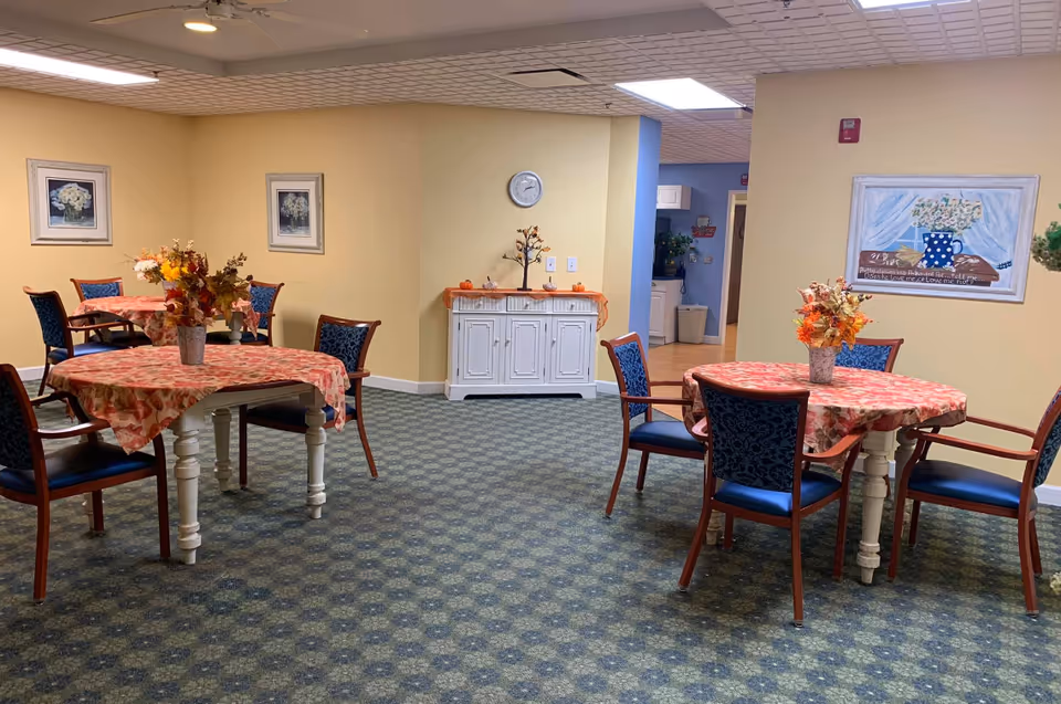 A dining area in a senior living facility with round tables covered in floral tablecloths and decorated with flower arrangements. The room has blue cushioned chairs with wooden frames, patterned carpet, and light yellow walls adorned with framed artwork. A white cabinet with small decorative pumpkins and a clock on the wall is visible in the background, along with a hallway leading to another room.