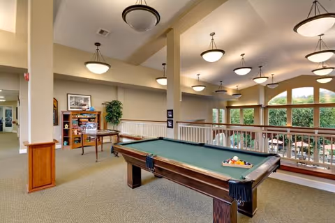 Interior view of a senior living facility's recreational area featuring a pool table in the foreground. The space has beige walls, carpeted floors, and multiple ceiling lights. There is a wooden bookshelf with various items and a potted plant against the wall. Large windows in the background provide natural light and a view of outdoor greenery.